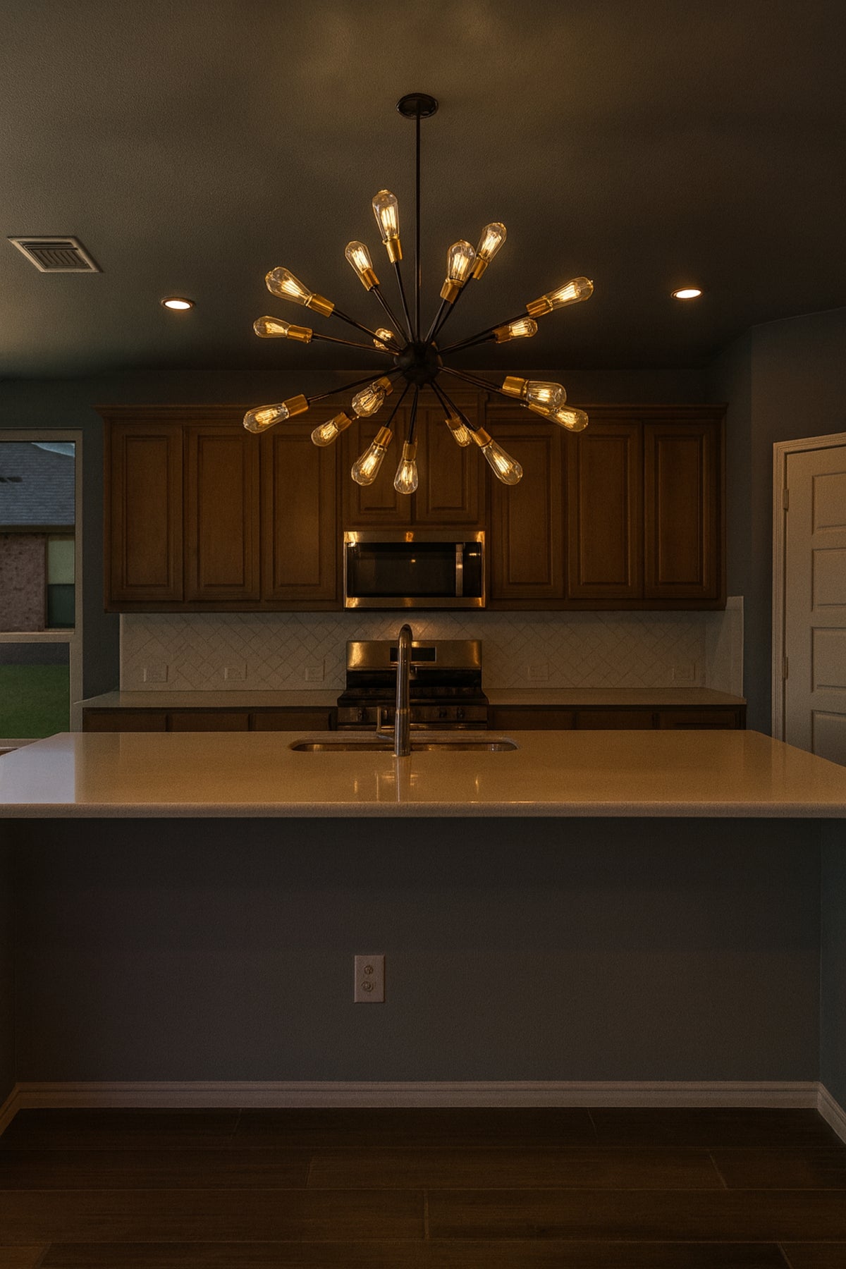 Modern kitchen with a large chandelier, dark walls, and wooden cabinets.